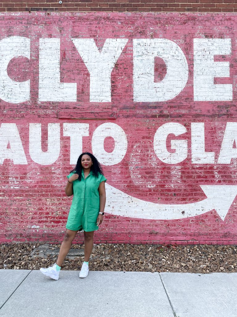 Kimberli is standing in front of a large, painted red wall with the words 'CLYDE AUTO GLASS' in bold white letters. She is wearing a green romper and white sneakers with green accents. She is standing with one foot forward and smiling while holding a small object in her left hand. The ground beneath her is a mix of gravel and concrete.
