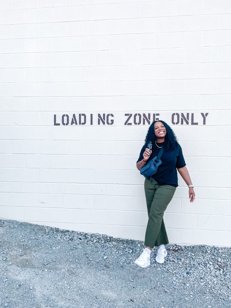 Kimberli smiling and standing against a white wall with the words 'LOADING ZONE ONLY' painted in bold letters. She is wearing a black t-shirt, olive green pants, and white sneakers. She has a blue fanny pack slung across her body and holds a small object in her hand. The ground beneath her is gravel.