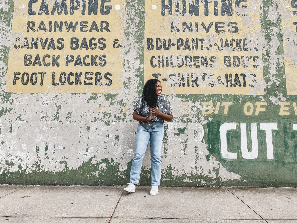 Kimberli standing in front of a weathered wall with vintage advertising text, including 'CAMPING,' 'RAINWEAR,' 'FOOT LOCKERS,' and other items. She is wearing a patterned gray shirt, blue jeans, and white sneakers, smiling while standing with one hand on her hip. The wall shows peeling paint, giving it a rustic, worn look.