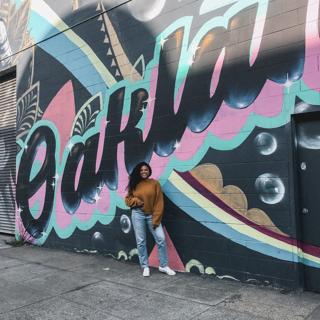Kimberli standing in front of a colorful mural that spells out 'Oakland' in bold, stylized lettering. The mural features a mix of pastel colors with swooping shapes and details like stars and bubbles. She is wearing a mustard yellow sweater, blue jeans, and white sneakers, smiling and posing in front of the vibrant artwork.