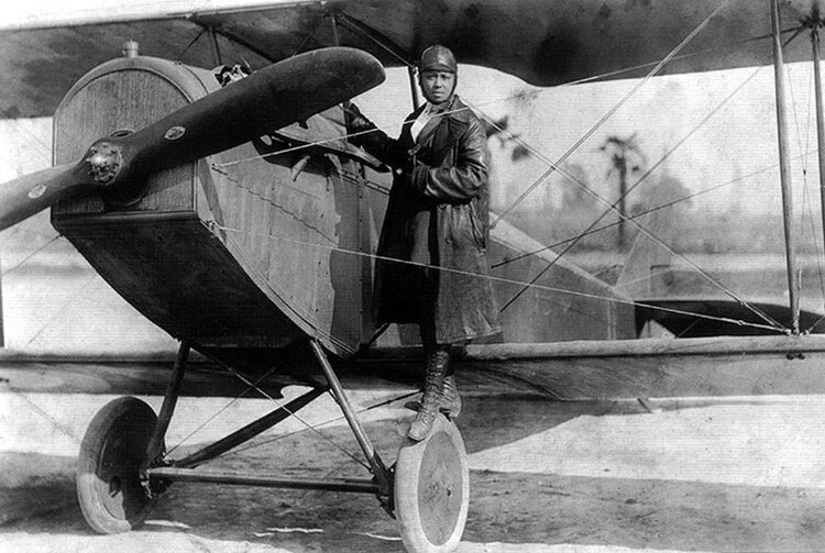 Black and white Bessie Coleman in a flying suit in front of an old fashioned airplane