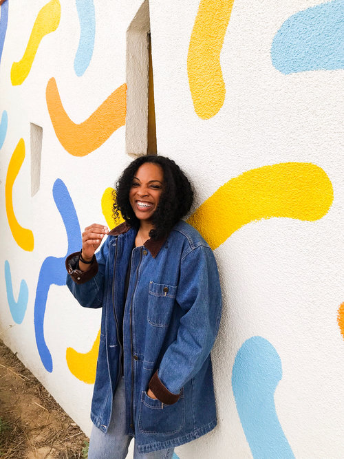 Kim standing in front of a wall smiling and wearing a long denim jacket with light colored jeans