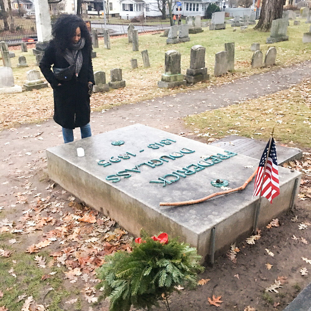 Kim standing in a long black coat with a black fanny pack across her check looking at Frederick Douglass’ gravestone in Rochester New York on a rainy day