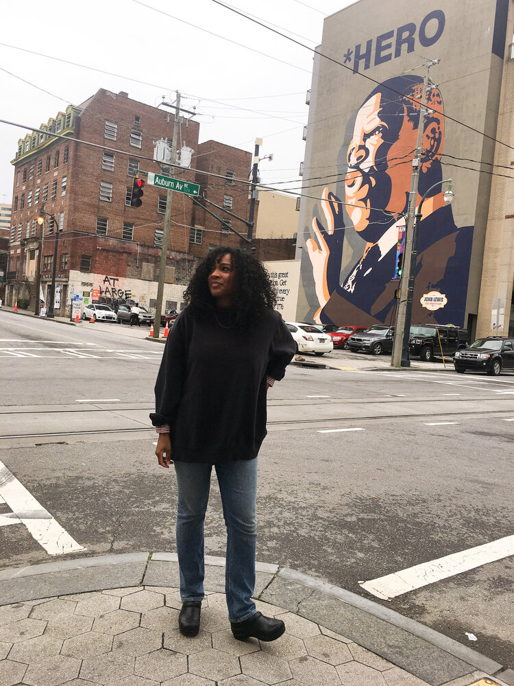 Kim standing with a large black sweater and blue jeans on the corner of Auburn Avenue with John Lewis Hero in the background in Atlanta Georgia