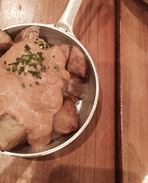 Metal bowl of hot patatas bravas with dark red brava sauce garnished with green chives