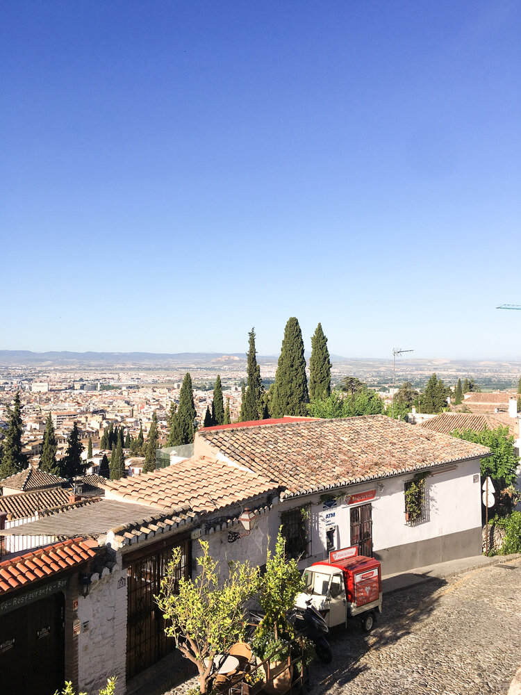 Small cobble stone street in the sun with views of the city of Granada and a parked red and white ice cream truck