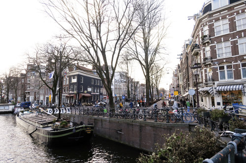 The canals in Amsterdam with bikes chained on the side and people walking on the sidewalk on a sunny afternoon