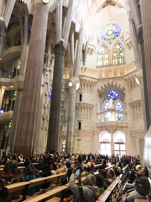 Tourists sitting in the pews inside the Sagrada Familia taking photos and looking up