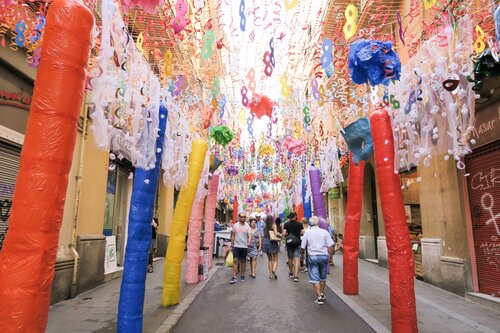 Tourists walking down a narrow street decorated with colorful ribbons and displays in the Gracia neighborhood