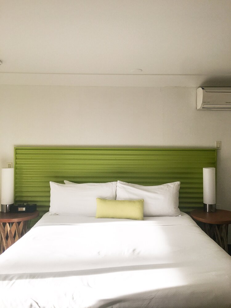 Hotel room with one bed made with white sheets and bright green headboard with the sun shinning in