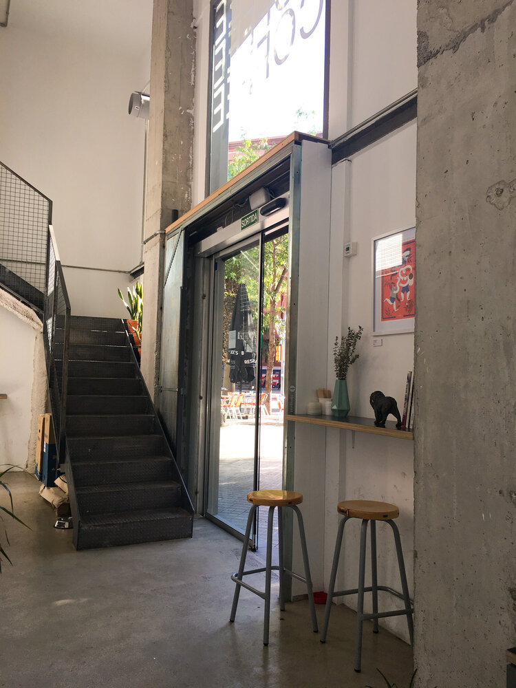 Inside the front entrance of Three Marks coffee shop in Barcelona with metal staircase and two wooden bar stools