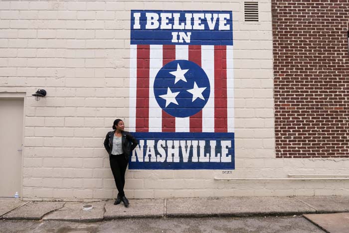 Kim standing in front of the "i believe in nashville" mural in nashville wearing black jeans, a green top, and a black leather jacket.