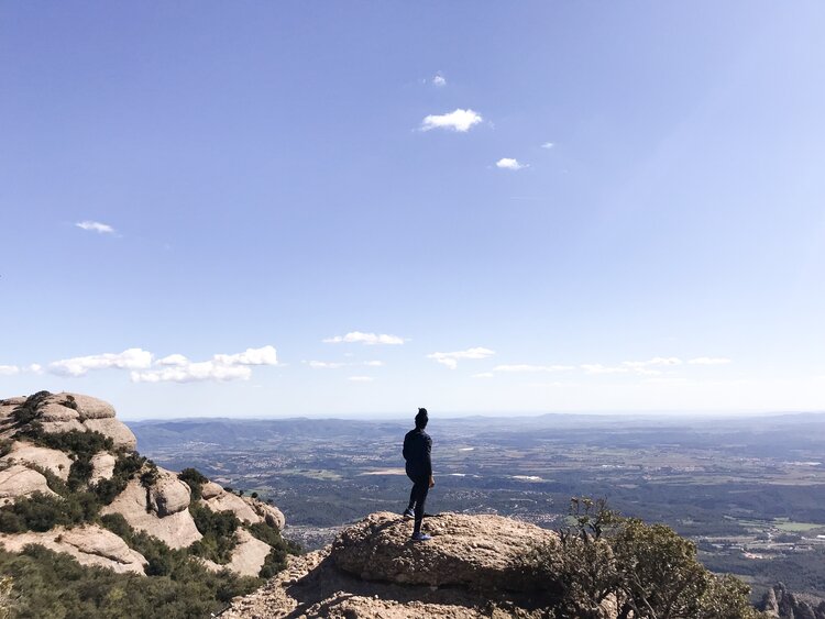 Kim traveling alone and standing on a giant rock at Montserrat looking out at the views of Barcelona on a sunny day
