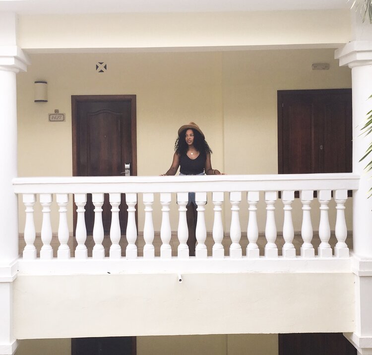 Kim traveling alone and looking out on a balcony wearing a beach hat and denim skirt and black tank top
