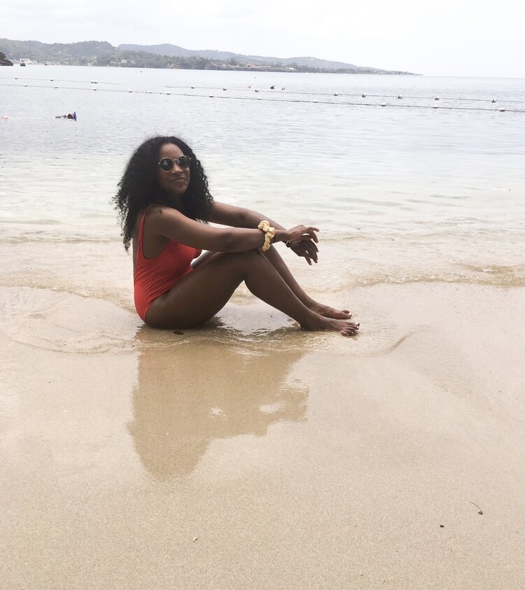 Kim traveling alone and sitting on the beach wearing a red one piece bathing suit with a yellow scrunchie around her wrist