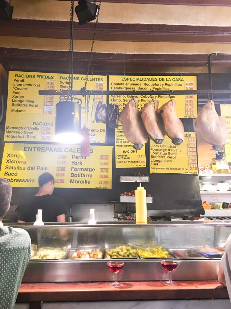 Large yellow menu on the wall and dried meat hanging from the ceiling with two glasses of rose cava inside one of the best tapas bars in Barcelona La Xampanyeria