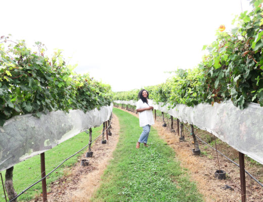Kim standing in a vineyard with a white dress and jeans on a bright and sunny day.