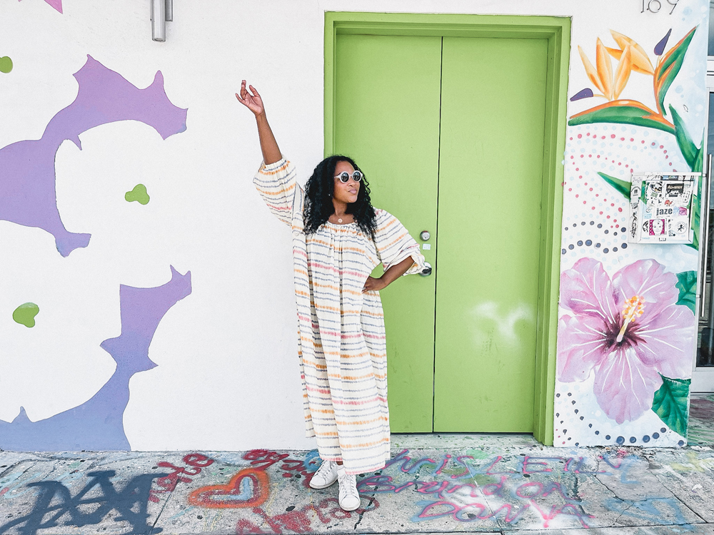 Kim standing in a colorful full length dress with sunglasses on a sunny day in Miami in front of a floral wall ready to travel alone.