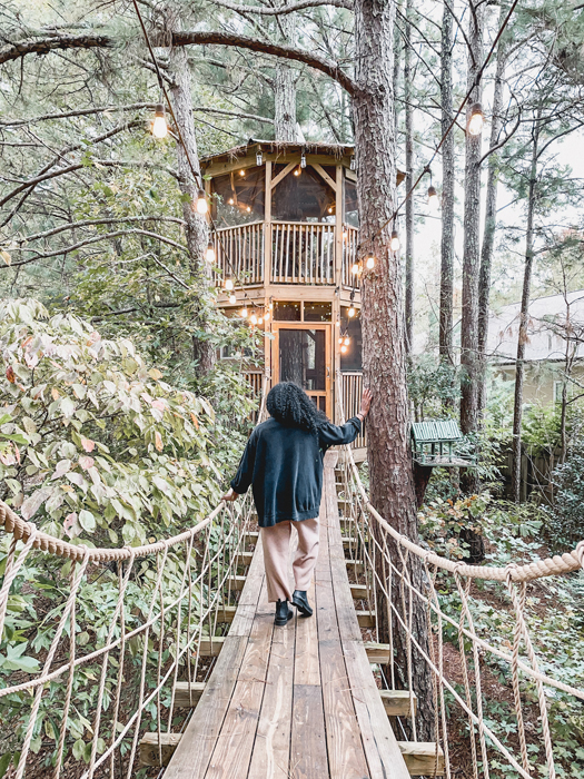 Kim standing on a bridge at a treehouse with twinkly lights on a sunny day