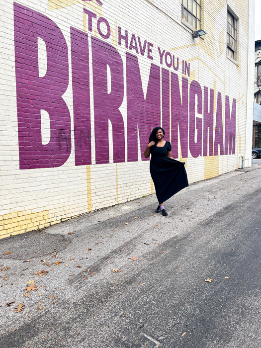 Kim standing in a full length dress in front of a wall mural that says Happy to have you in Birmingham while traveling alone